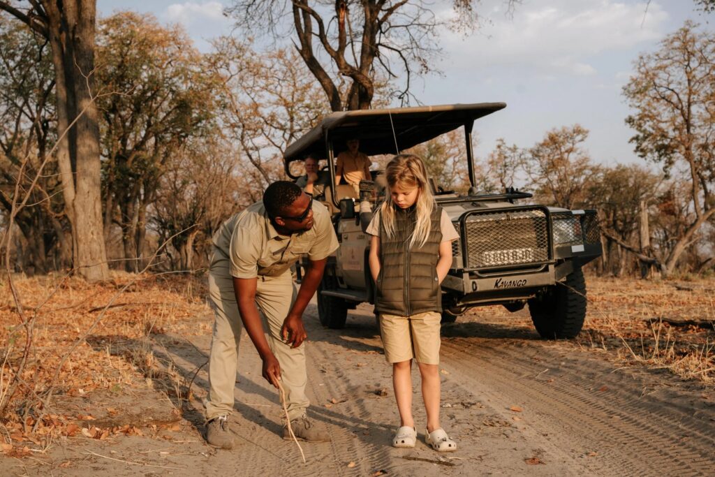 En mann viser en ung jente dyrespor på en støvete vei, med en safarijeep i bakgrunnen